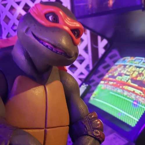 A close-up of a turtle figurine wearing a red mask, with a playful expression, set against a colorful arcade game background.