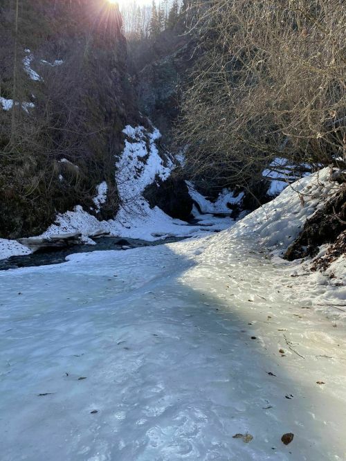 A serene winter landscape featuring a frozen river surrounded by snow-covered banks and trees. Sunlight peeks through.
