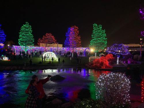 Colorful holiday lights illuminate trees and decorations around a pond, with people enjoying the festive atmosphere at night.