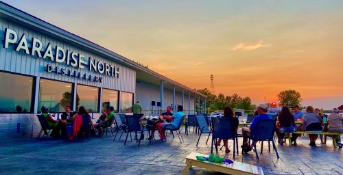People relax on a patio at Paradise North Distillery, enjoying a sunset view and socializing.