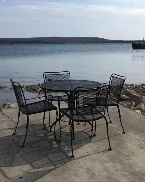 A black metal table with four chairs by a calm lake, surrounded by rocks and distant hills under a clear sky.