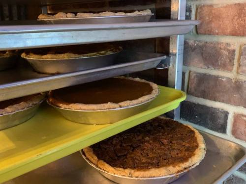 Pies cooling on metal shelves, including pumpkin and pecan varieties, against a brick wall background.