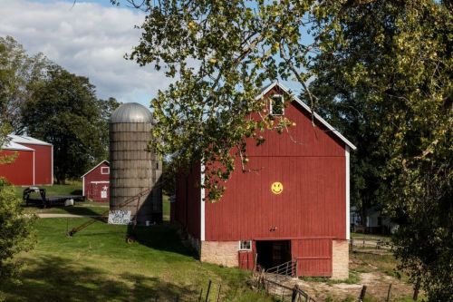 A red barn with a smiley face, a silo, and green trees under a partly cloudy sky.
