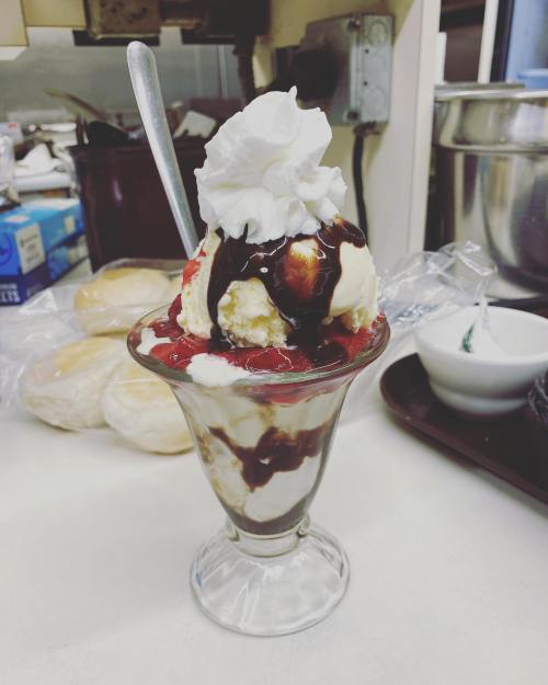 A delicious sundae in a glass dish, topped with whipped cream, chocolate syrup, and cherries, with rolls in the background.
