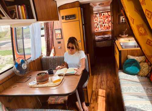 A woman sits at a table in a cozy camper, reading a book with a plant and fan nearby, sunlight streaming in.