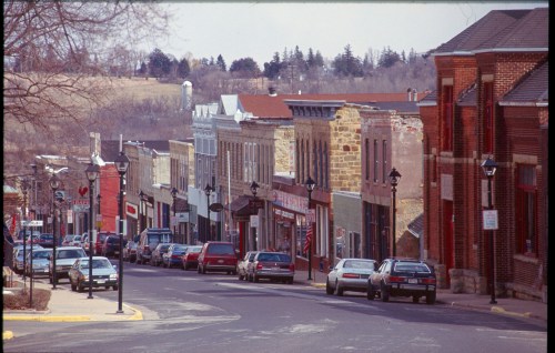 A quaint street lined with historic buildings and parked cars, set against a backdrop of rolling hills.