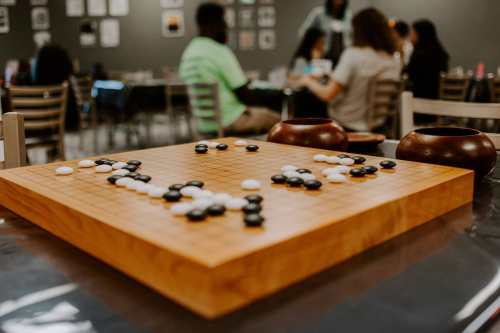 A wooden Go board with black and white stones, set in a busy room with people playing and socializing in the background.