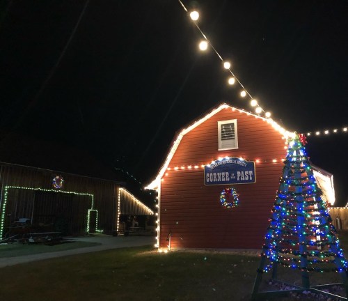 A red barn illuminated with festive lights at night, featuring a decorated Christmas tree and a sign reading "Corner of the Past."