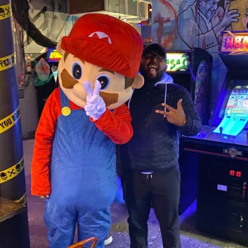 A person poses with a large Mario mascot in an arcade, both smiling and having fun. Colorful arcade games in the background.