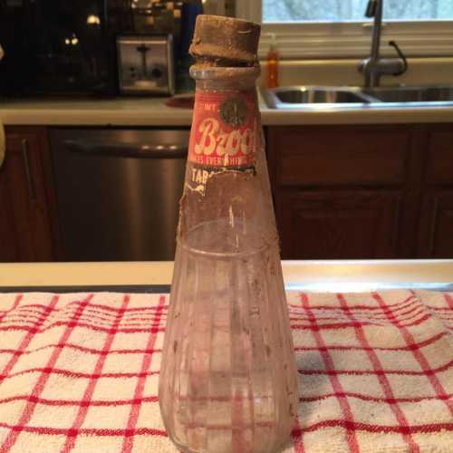 A vintage glass bottle with a cork stopper and a faded label, resting on a red and white checkered cloth.