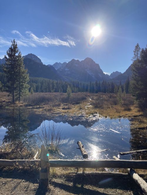 A serene landscape featuring a reflective pond, mountains in the background, and a bright sun shining in a clear blue sky.