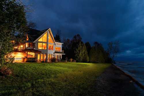 A lakeside house illuminated at night, surrounded by trees and a dark, cloudy sky.