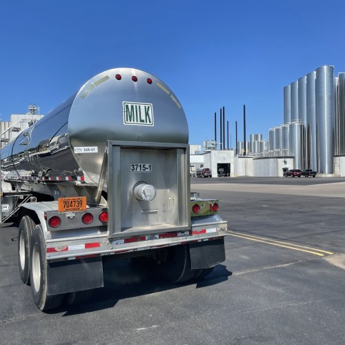 A milk tanker truck parked in an industrial area with large silos and buildings in the background.