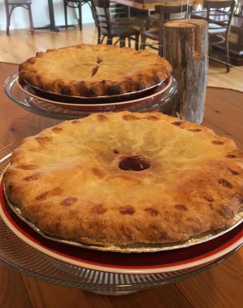Two golden-brown pies on glass stands, with a rustic wooden table and chairs in the background.