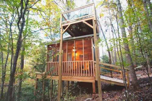 A wooden treehouse nestled among trees, featuring a balcony and warm lighting, surrounded by lush greenery.