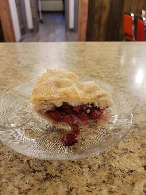 A slice of cherry pie on a glass plate, with a crumbly crust and cherry filling, on a countertop.