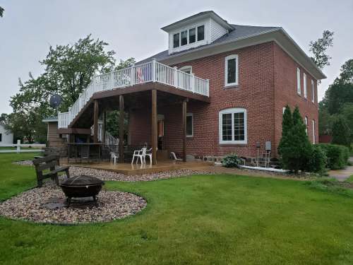 A two-story brick house with a large deck, surrounded by green grass and trees, featuring a fire pit in the yard.