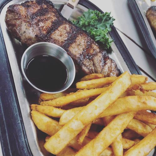 A grilled steak on a metal plate with a side of crispy fries and a small cup of dipping sauce.