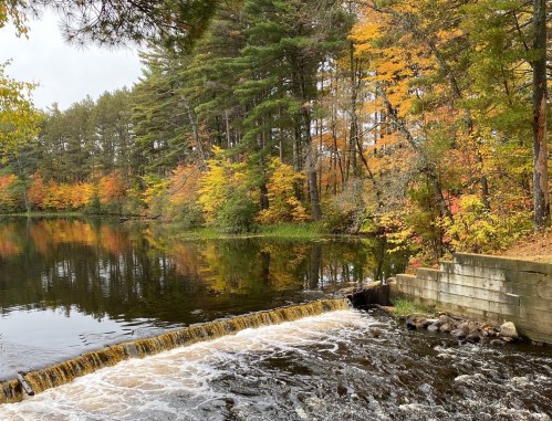 A serene river scene with autumn foliage reflecting on the water, featuring a small waterfall and lush trees.