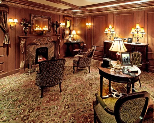 Cozy, elegantly furnished room with a fireplace, ornate chairs, and warm lighting, featuring rich wood paneling and decor.