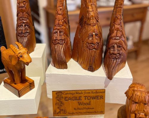 Wooden carvings of eagle towers and a goat figure displayed on shelves, with a sign detailing the artist and materials used.