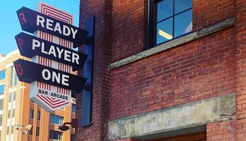 Sign for "Ready Player One" bar-arcade on a brick building, featuring retro-style lettering against a blue sky.