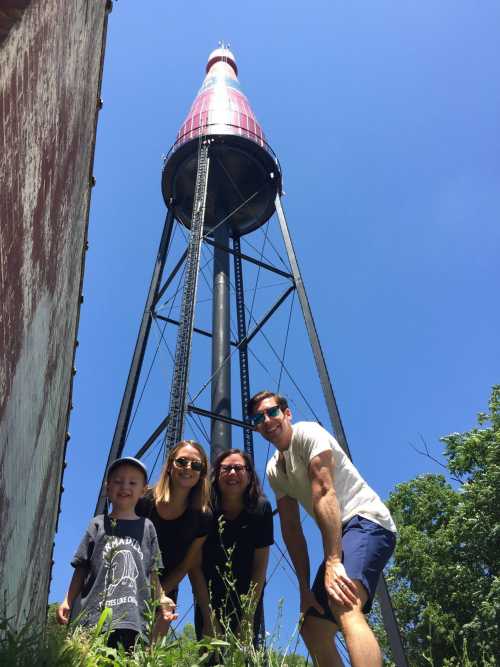 A family of four poses in front of a tall, red and white lighthouse against a clear blue sky.