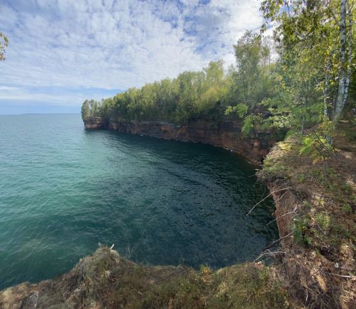 A serene view of a rocky shoreline with lush greenery and calm blue waters under a partly cloudy sky.