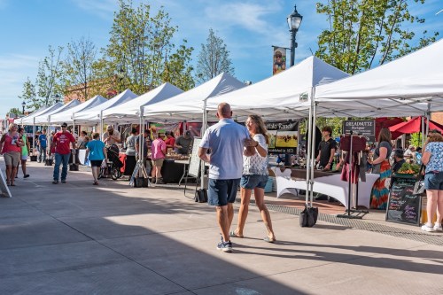 A bustling outdoor market with white tents, vendors, and visitors enjoying the sunny day.