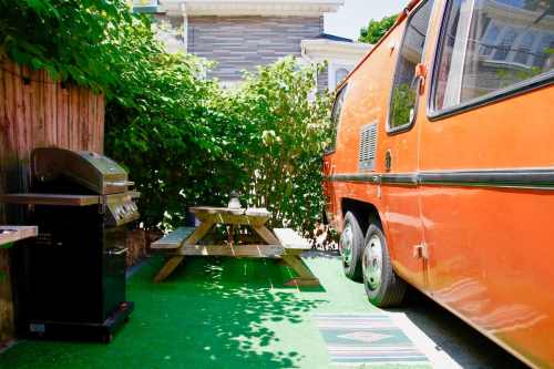 An orange trailer parked beside a picnic table and grill, surrounded by greenery in a cozy outdoor space.