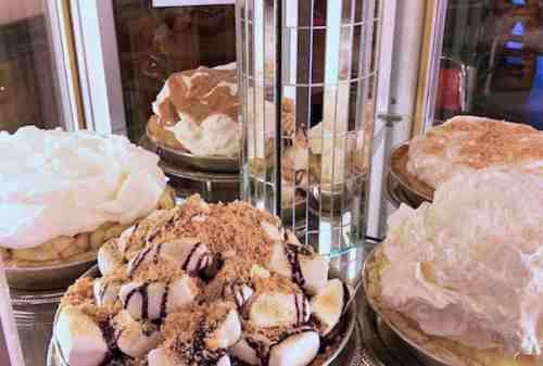 A display case filled with various desserts, including cream pies and a layered cake topped with whipped cream and crumbs.