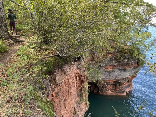 A person walks along a wooded path beside a rocky cliff overlooking a calm blue lake.