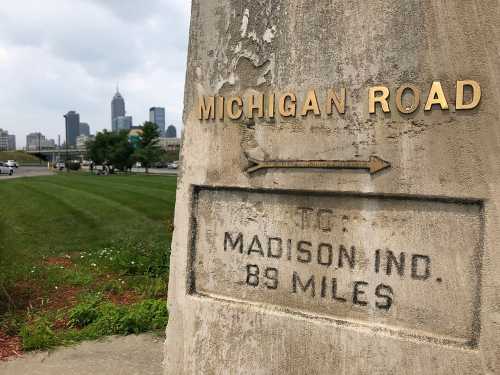 Signpost for Michigan Road, indicating distance to Madison, Indiana, with a city skyline in the background.