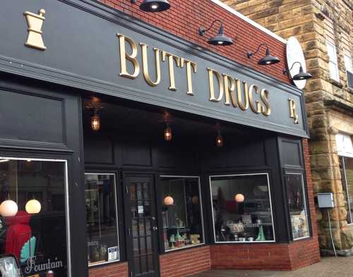 A storefront with the sign "BUTT DRUGS" prominently displayed, featuring large windows and decorative lighting.