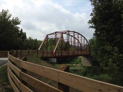 A red metal bridge spans a river, surrounded by trees and a wooden fence along a pathway.