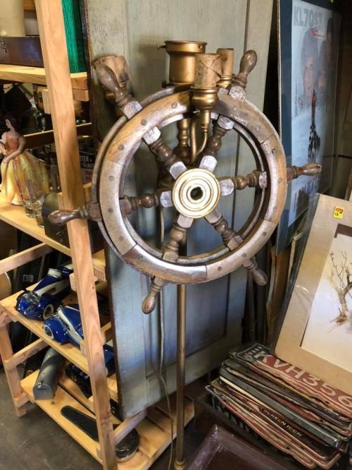 A vintage ship's wheel displayed on a stand, surrounded by various antique items in a shop.