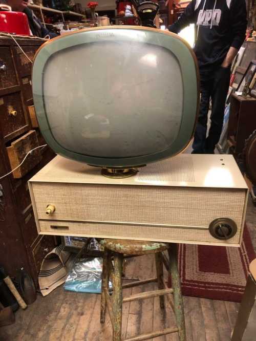 A vintage television set with a round screen and a wooden base, displayed on a stool in a cluttered room.