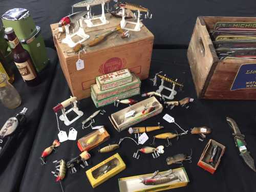 A collection of vintage fishing lures and tackle displayed on a table, alongside wooden boxes and bottles.