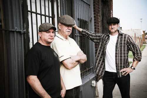 Three men in flat caps stand against a brick wall, with one leaning on a fence, exuding a casual, confident vibe.