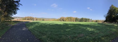 A panoramic view of a green field bordered by trees under a clear blue sky with a few clouds.