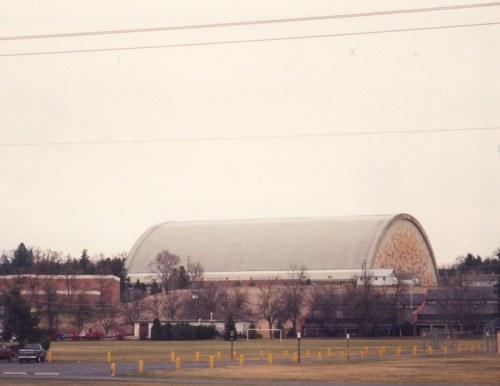 A large, curved building with a yellow and gray roof, surrounded by trees and a grassy area. Overcast sky above.