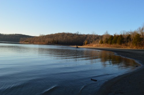 A serene lakeside view with calm water, sandy shore, and trees lining the distant hills under a clear blue sky.