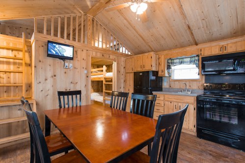 Cozy wooden cabin interior featuring a dining table, kitchen appliances, and a lofted sleeping area.