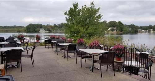 Outdoor dining area with empty tables, surrounded by flowers, overlooking a calm lake under a cloudy sky.