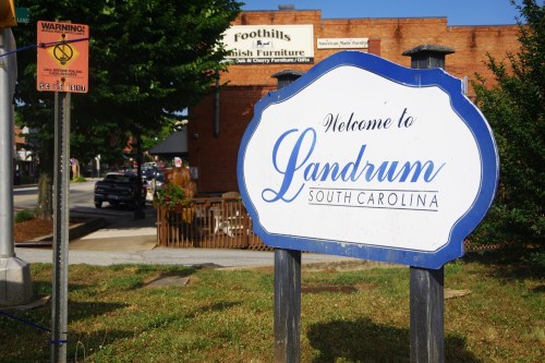 Welcome sign for Landrum, South Carolina, with nearby buildings and a warning sign visible in the background.