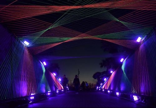 A colorful illuminated bridge at night, featuring vibrant strings of light and silhouettes of people walking through.
