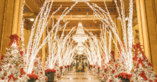 A festive hallway adorned with white lights, Christmas trees, and red bows, leading to a chandelier.