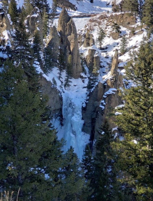 A frozen waterfall surrounded by snow-covered rocky cliffs and evergreen trees in a winter landscape.