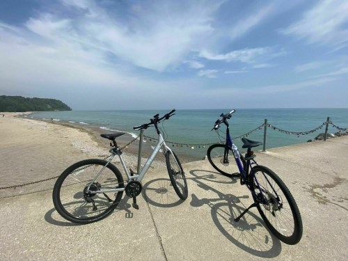 Two bicycles parked on a concrete path by the beach, with calm water and a cloudy sky in the background.