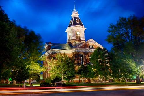 Historic building illuminated at night, surrounded by trees and a blue sky, with light trails from passing vehicles.